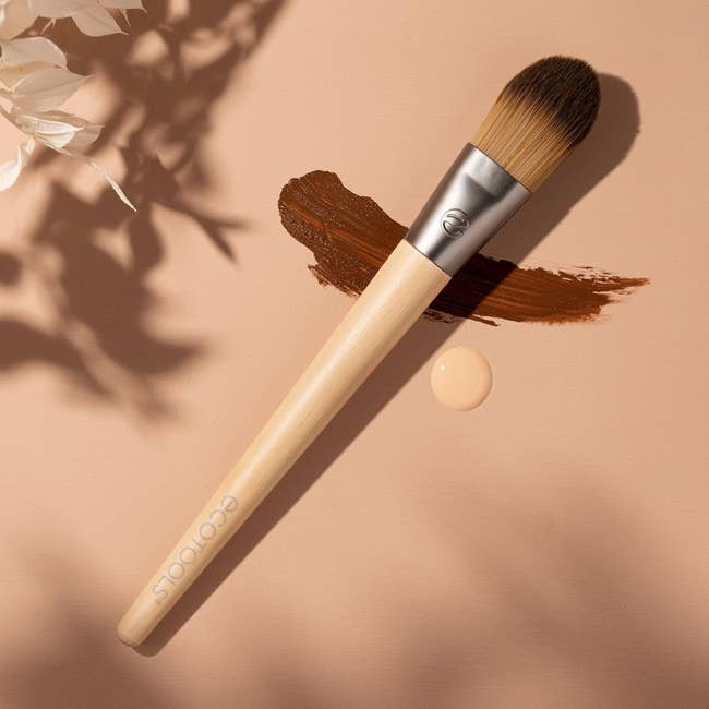 Makeup brush with wooden handle placed on a surface next to a makeup smear and a drop, with decorative leaves nearby