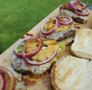 Burgers topped with grilled pineapple, reddish onion, and lettuce connected a woody board, with bare buns to the side