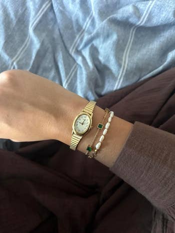 Person's wrist with a gold watch and layered bracelets, on a fabric surface