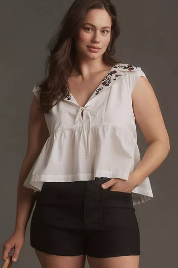 Woman in a ruffled blouse with embroidery and black shorts, posing with a hand on hip