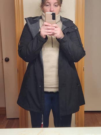 Person in a mirror selfie wearing a layered outfit with a black coat over a beige sweater