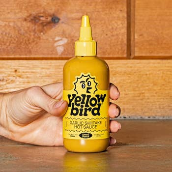 Social media trends Viral content Trending hashtags Social media updates Trending memes and videos Hand holding a bottle of Yellowbird Garlic Shiitake Hot Sauce in a kitchen setting, front label visible