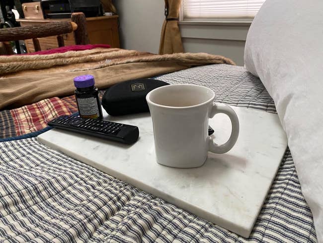 A marble cutting board tray with a mug, remote, and container on a bed