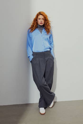 Person in a blue shirt, layered over a black turtleneck, paired with wide-leg grey trousers and white sneakers