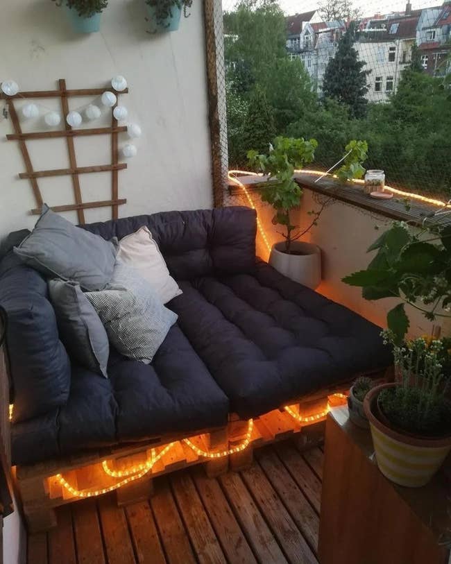 Black tufted cushions and gray throw pillows on top of wooden pallets with twinkle lights inside on a wooden balcony
