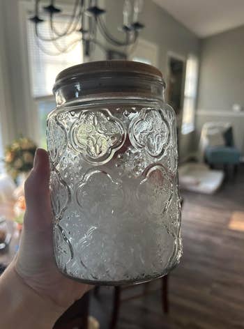 Person holding a decorative glass jar with a wooden lid in a well-lit room. Room appears to have a wooden floor, a chandelier, and furniture in the background