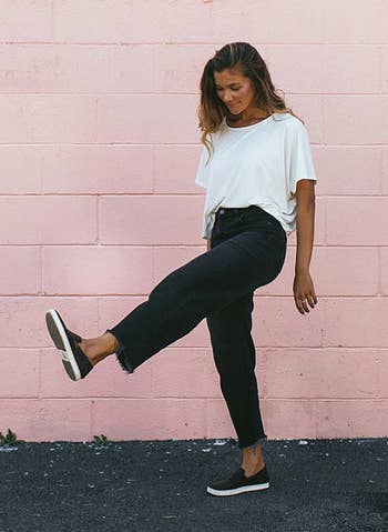 Model wearing the sneakers in black in front of a pink wall kicking their feet