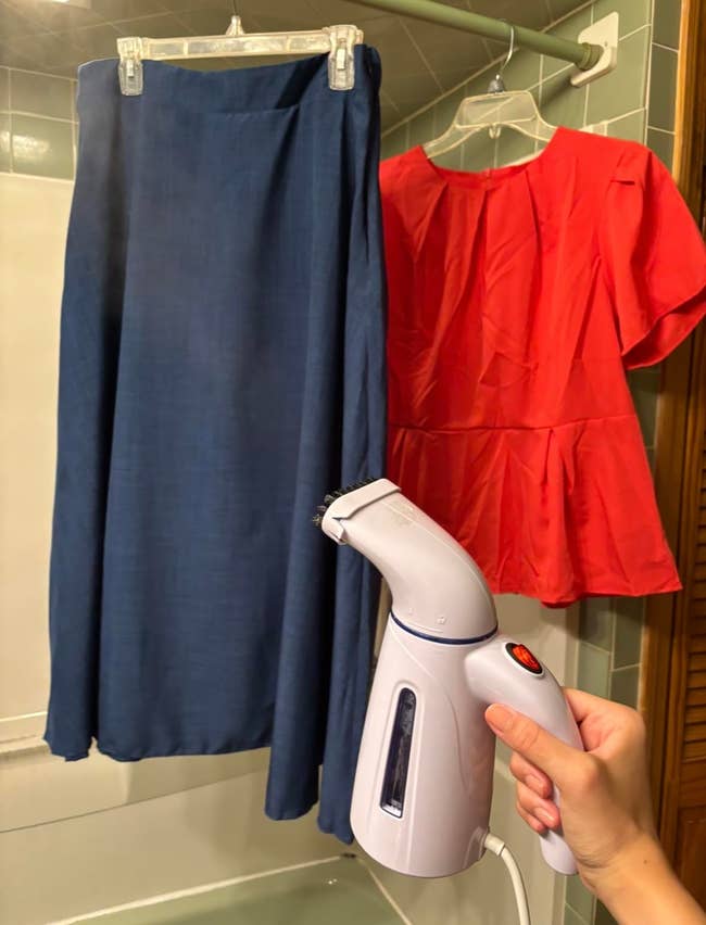 Hand holds a garment steamer adjacent to a bluish skirt and reddish apical hanging successful a bathroom
