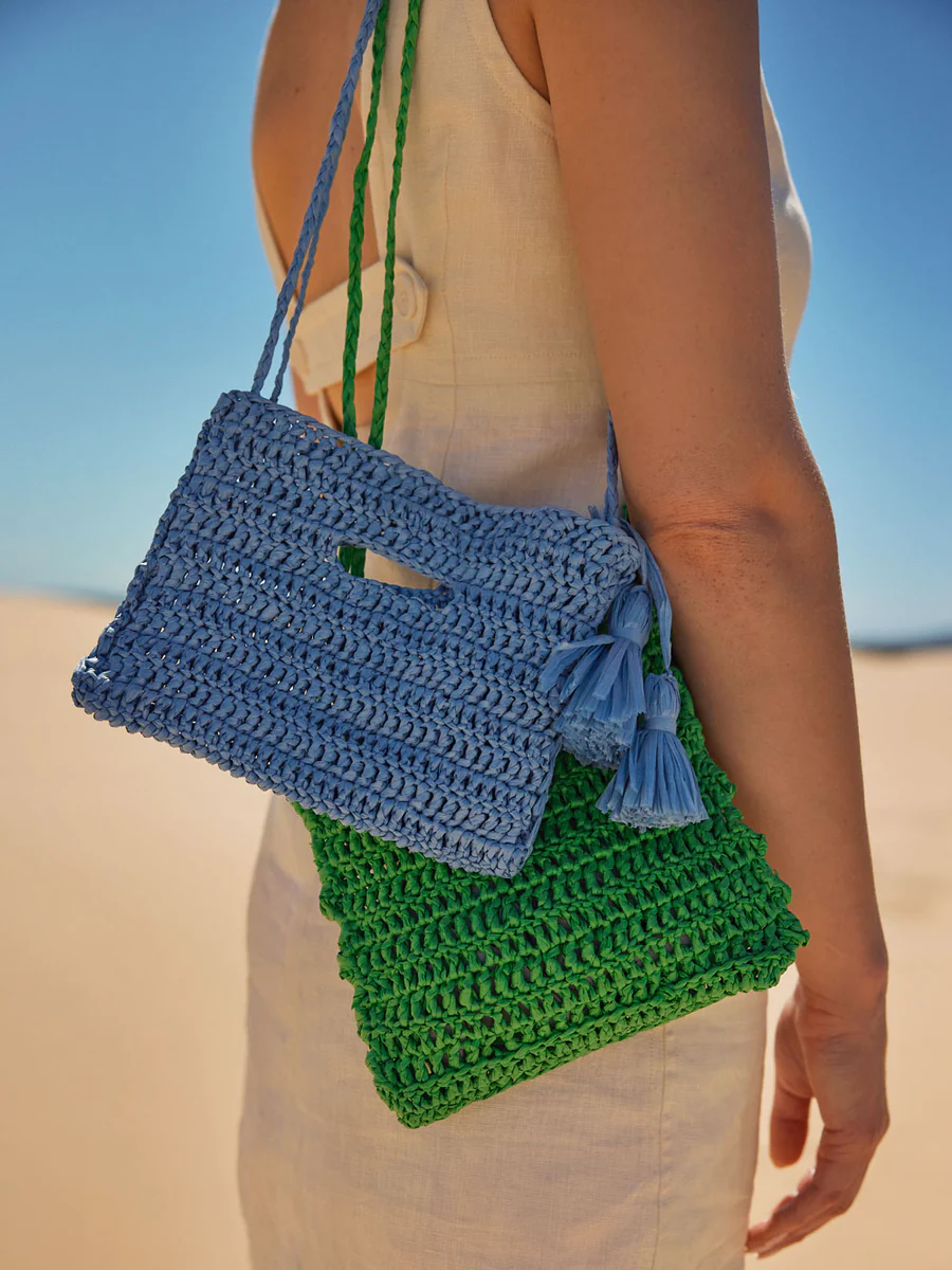 Person holding two hand-knitted bags, one green and one blue, with tassel detail, against a desert background