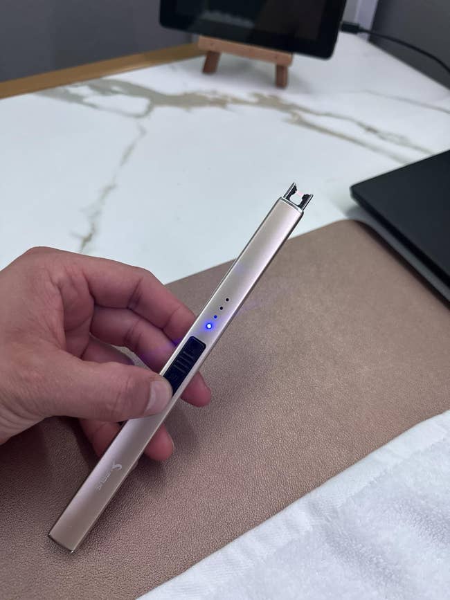 A manus  holds a sleek electrical  lighter with a disposable   bluish  airy  indicator, resting connected  a table  adjacent  to a laptop