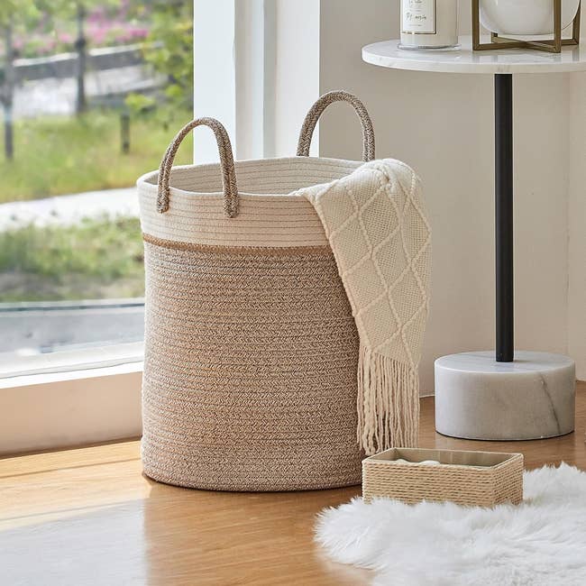 Large woven handbasket with handles, holding a fringed blanket, beside a model and tiny textured box. Ideal for stylish location storage
