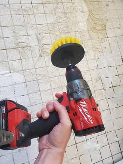 Hand holding a red cordless drill with a yellow scrubbing brush attachment, cleaning grout on tiled wall