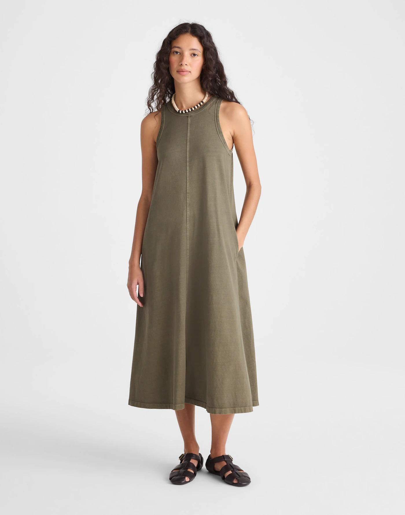 Person wearing an olive-green jersey midi tank dress with fisherman sandals, standing with one hand in pocket