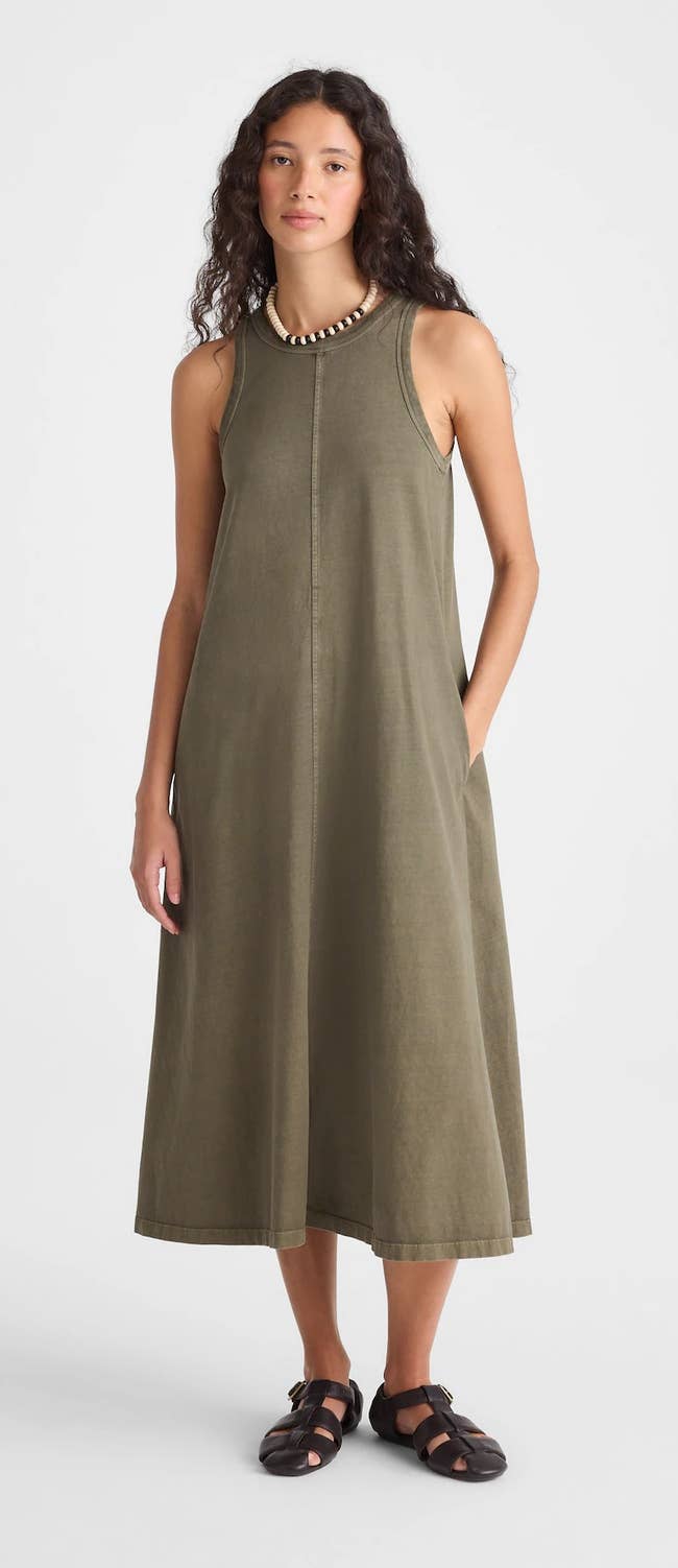 Person wearing an olive-green jersey midi tank dress with fisherman sandals, standing with one hand in pocket