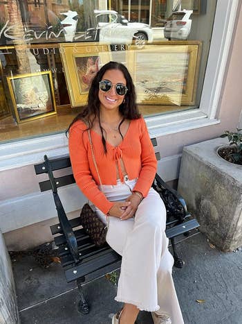 Person sitting on a bench outdoors, wearing a stylish long-sleeve top and wide-leg pants, accessorized with sunglasses and a handbag