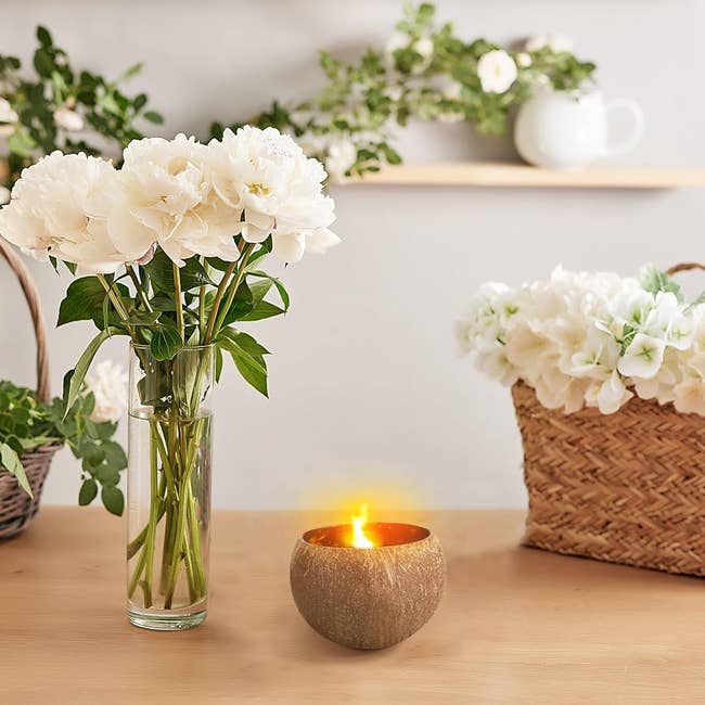 A vase with white peonies on a table next to a lit candle, with a basket of flowers in the background. Perfect for home decor shopping