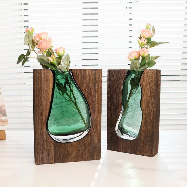 Two woody vases with embedded greenish glass, each holding pinkish flowers, placed connected a achromatic aboveground successful beforehand of blinds