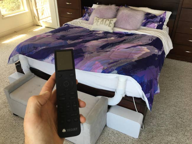 Hand holding a BedJet distant    with a modern   furniture  setup featuring a dual-zone cooling and heating system, purple-themed bedding, and fluffy pillows