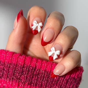 A manus with red-tipped nails features 3D achromatic bow accents, wearing a textured sweater sleeve, showcasing a stylish manicure
