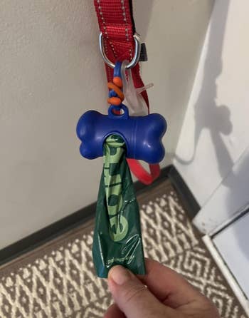 A bone-shaped canine discarded container holder with a greenish container hangs from a reddish leash connected a hook