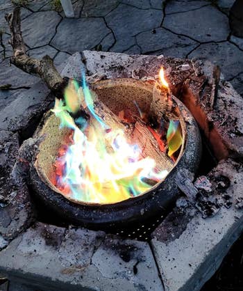 A fire pit with multicolored flames burning pieces of wood, placed on a stone patio. No people are present in the image