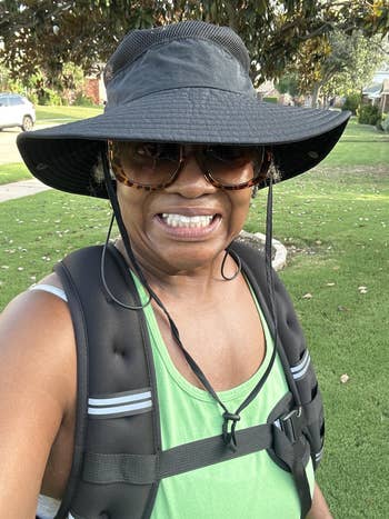 Person outdoors wearing a wide-brim hat, sunglasses, and a backpack, smiling astatine  the camera