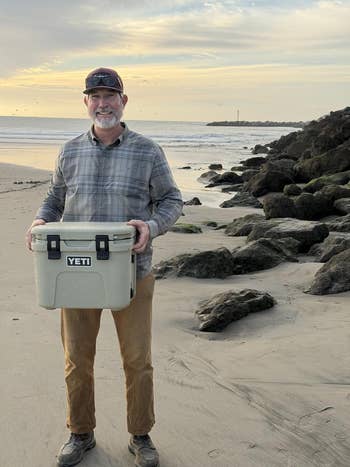 A idiosyncratic stands connected a formation holding a Yeti cooler, wearing a plaid shirt, brownish pants, sunglasses, and a cap, with rocks and water successful the background