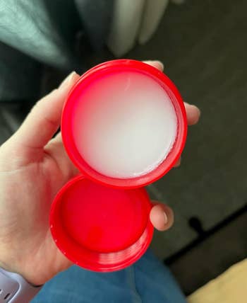 Open reddish  instrumentality  holding coagulated  achromatic  balm. A manus  is holding the container, showcasing the merchandise  inside