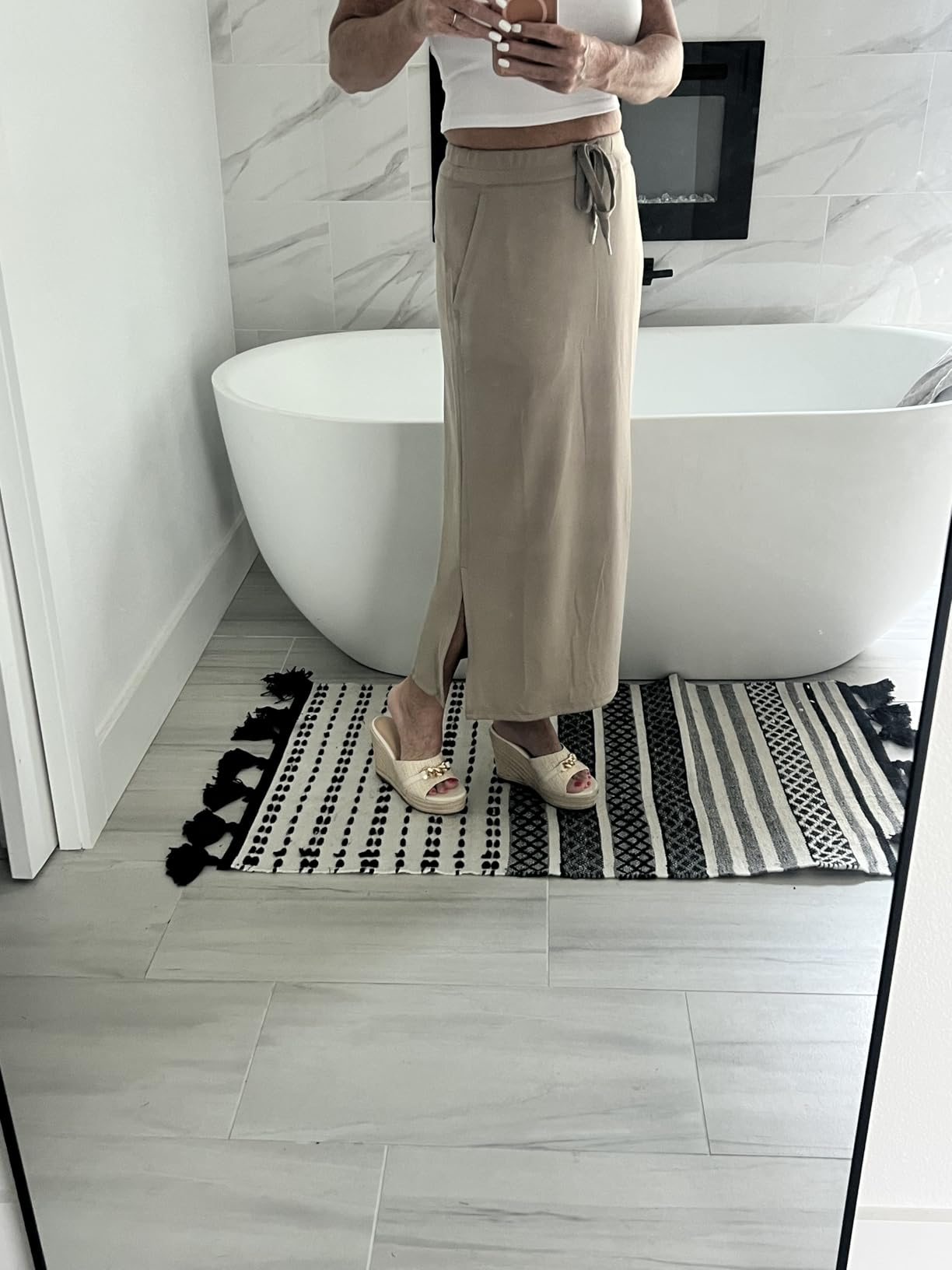 Person successful  a achromatic  vessel  apical  and beige maxi skirt with a tie, lasting  by a bathtub connected  a patterned rug, wearing wedge sandals