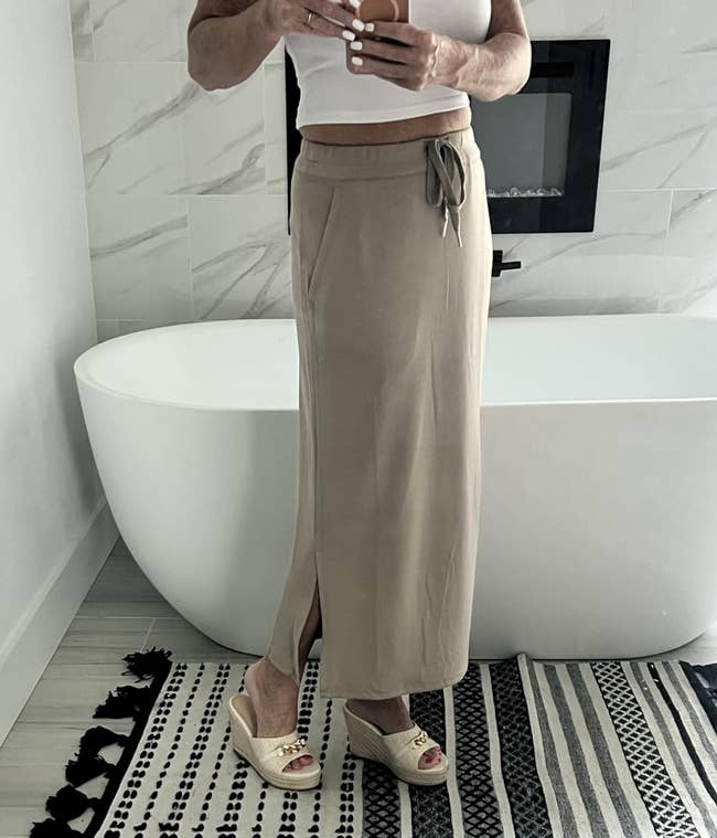 Person successful a achromatic vessel apical and beige maxi skirt with a tie, lasting by a bathtub connected a patterned rug, wearing wedge sandals