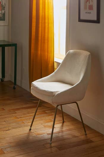 A modern cream-colored chair with sleek metal legs in a sunlit room with wooden floors, near golden curtains