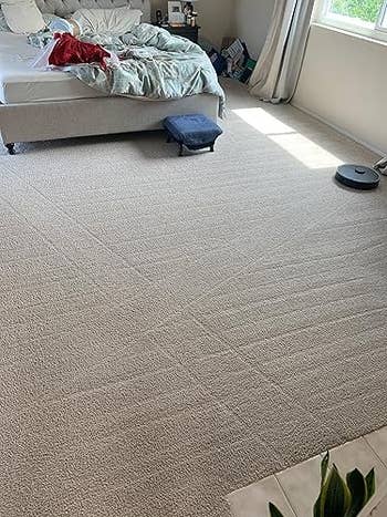 Bedroom with unmade bed, robotic vacuum, and model   light. Carpet covers the floor, with a tiny  ottoman nearby