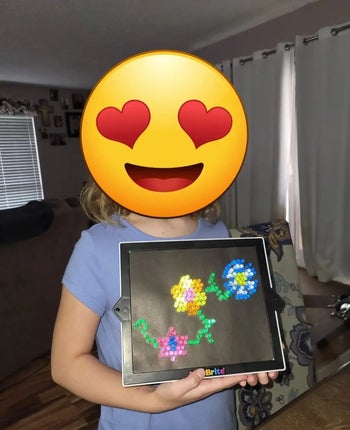 reviewer photo of their kid holding a Lite-Brite with a flower design on it and a heart-eye emoji covering their face