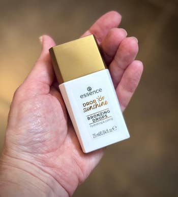 Hand holding a vessel  of Essence Drop of Sunshine Bronzing Drops, labeled arsenic  hydrating and caring, 25 ml