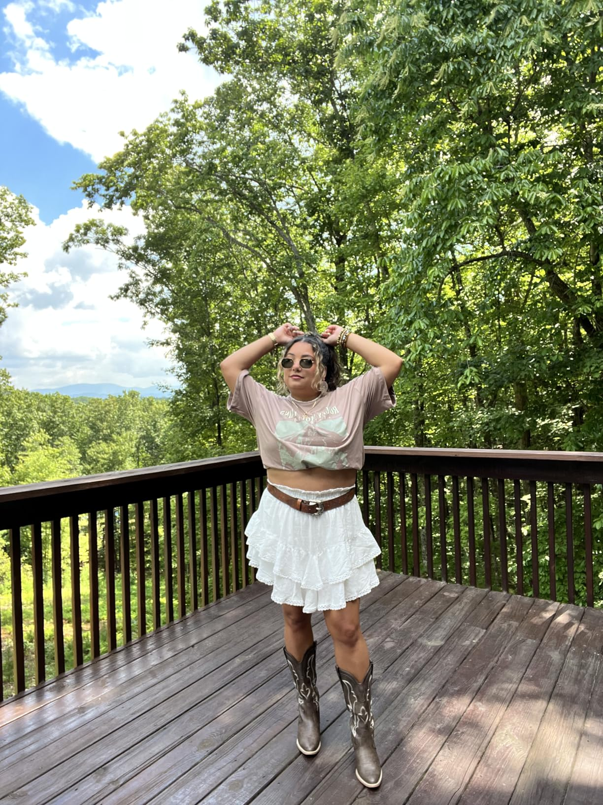 Person connected  a platform  wearing a graphic t-shirt, layered ruffle skirt, and cowboy boots, with wood  background