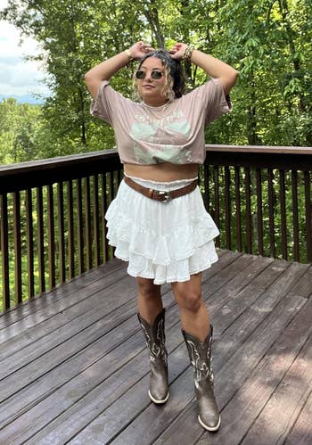 Person connected a platform wearing a graphic t-shirt, layered ruffle skirt, and cowboy boots, with wood background