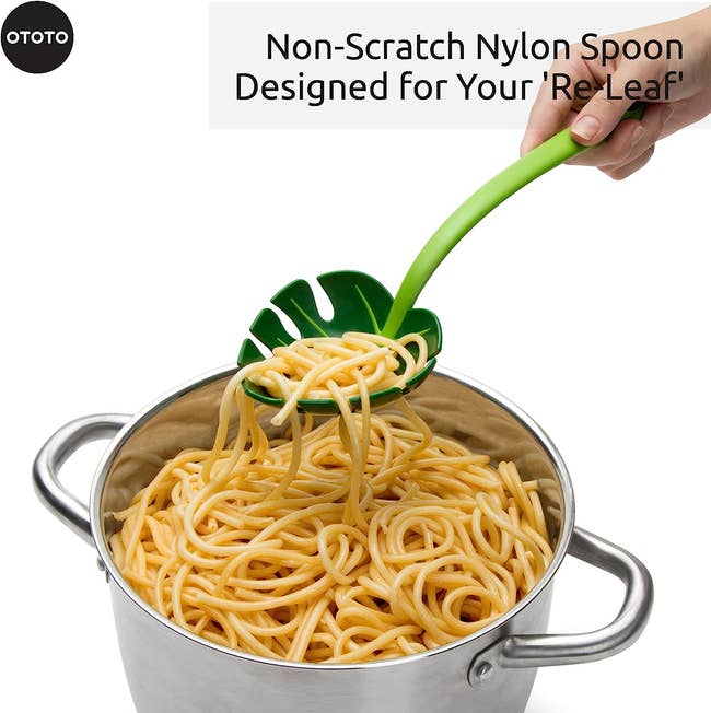 Hand holding a green nylon spoon shaped like a leaf, lifting spaghetti from a pot