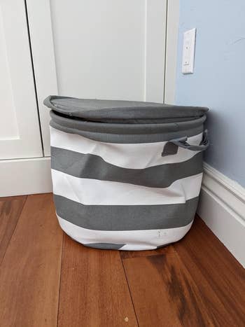 Collapsible laundry handbasket  with grey  and achromatic  stripes, placed connected  a woody  level  adjacent   a closed achromatic  door