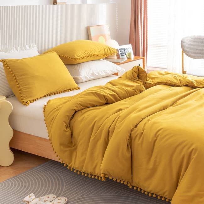Yellow comforter with pom pom fringe edging and matching pillows on white sheets on a light wooden bed frame