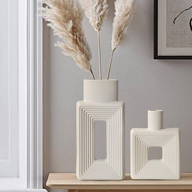 Two modern geometric ceramic vases with pampas grass on a wooden shelf beside a framed art piece