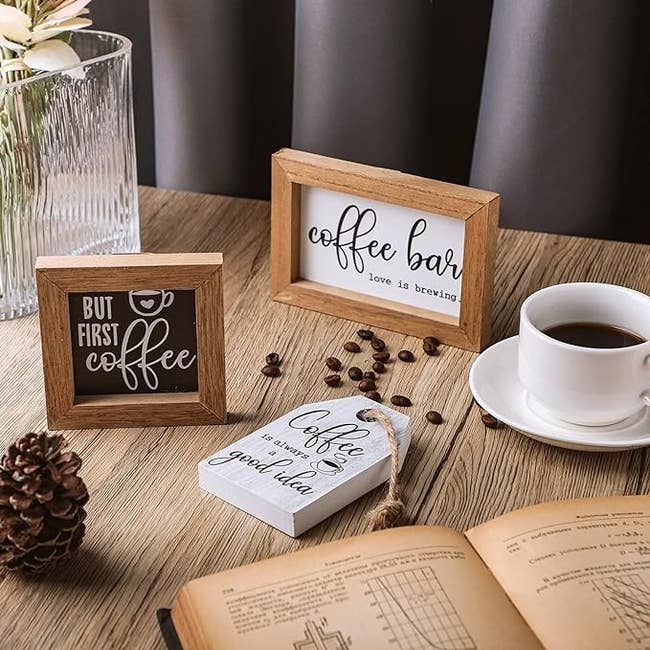 Wood-framed signs with coffee-related phrases beside a cup and saucer on a table with scattered coffee beans and an open book