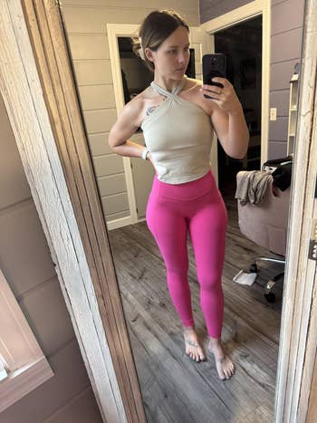 Person wearing a cross-strapped top and vibrant leggings, standing barefoot in a mirror selfie inside a room with wooden floors