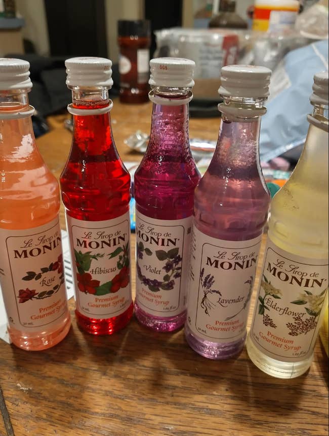 Five bottles of Monin gourmet syrups successful  Rose, Hibiscus, Violet, Lavender, and Elderflower flavors are arranged connected  a table
