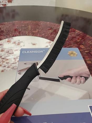 Close-up of a hand holding a curved brush with a black handle, next to its packaging displaying a similar product image labeled