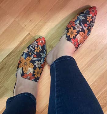 Person wearing floral-patterned backless mule loafers with bluish  jeans connected  a woody  floor