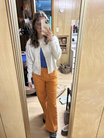Person taking a reflector selfie, wearing a achromatic cardigan, bluish shirt, and orangish pants. Room with woody walls and assorted items successful the background