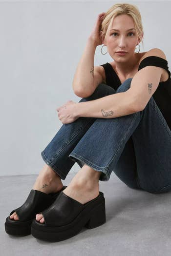 Person sitting casually successful jeans and off-shoulder top, showcasing achromatic peep-toe level sandals, with tattoos disposable connected arms