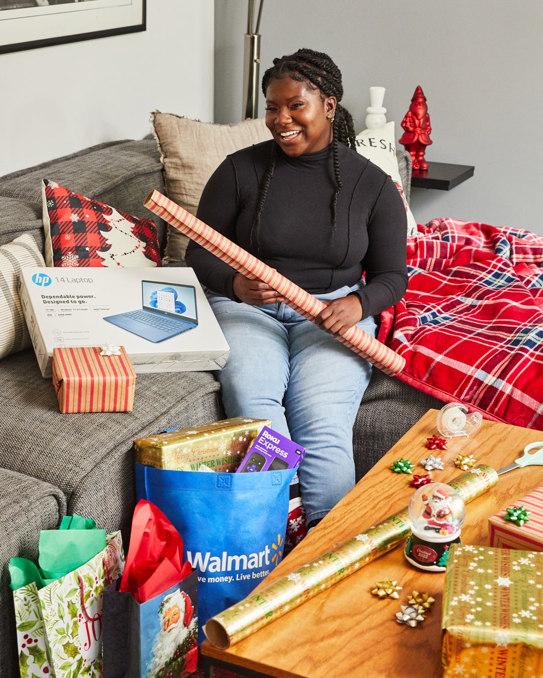 Elise sits on couch with wrapping paper in hands next to HP laptop, Walmart bag with Roku Express in it, and mini bows