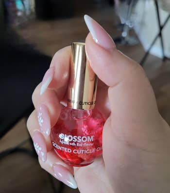 A person's hand with manicured nails holds a bottle of Blossom scented cuticle oil