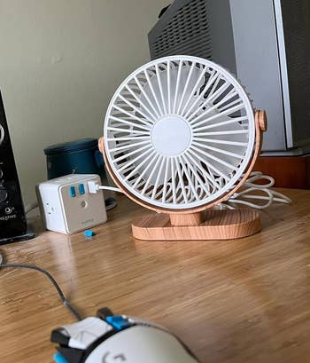 Small white and wood table fan on a wooden desk, next to a surge protector and a computer mouse